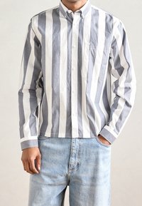 Homme portant une chemise à manches longues boutonnée avec de larges rayures verticales blanches et grises et un jean bleu clair, une main dans la poche.