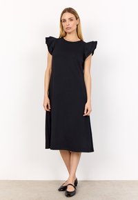 Eine Frau steht in einem schwarzen Midikleid mit rüschenbesetzten kurzen Ärmeln und schwarzen Mary Jane Schuhen vor einem schlichten weißen Hintergrund.