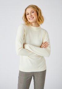 Maglione crema con scollatura rotonda a coste e maniche lunghe, realizzato in tessuto morbido. Abbinato a pantaloni a quadri con motivo.