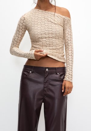 Femme portant un top beige en dentelle à une épaule avec manches longues et un pantalon en cuir marron foncé taille haute, main soulevant légèrement le top.
