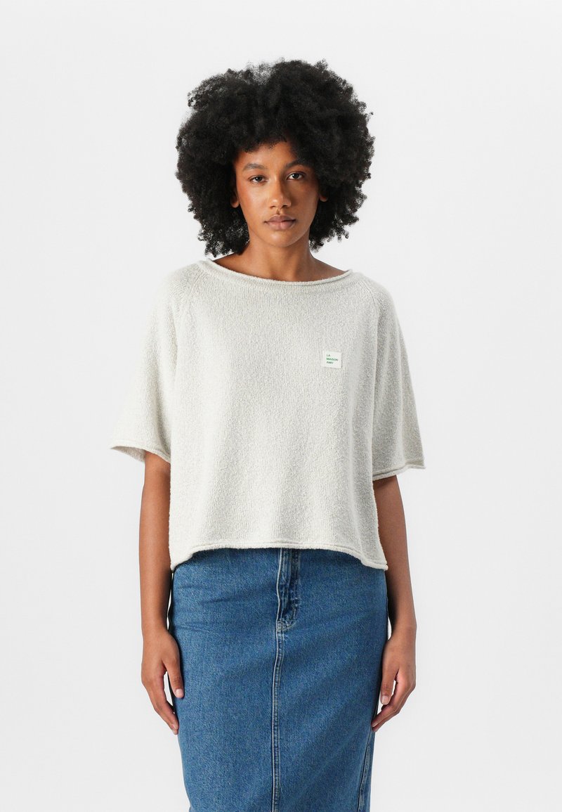 Light grey, short-sleeved top with a textured fabric and wide neckline. Features a small green label on the left chest. Paired with a denim skirt.