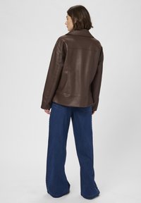Donna che indossa una giacca di pelle marrone e jeans a gamba larga blu, in piedi con la schiena rivolta verso la macchina fotografica su uno sfondo neutro.