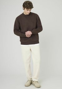 Pull marron avec une finition texturée, col rond et poignets côtelés, associé à un pantalon couleur crème et des chaussures beige clair.