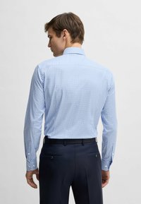 Niebiesko-biała koszula w kratę z długim rękawem i tradycyjnym kołnierzykiem, uszyta z materiału o gładkiej fakturze. Widoczne ciemne spodnie.