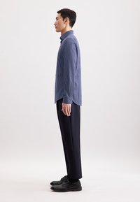 Uomo in profilo che indossa una camicia azzurra a fantasia, pantaloni neri e scarpe eleganti nere, su uno sfondo chiaro e semplice.