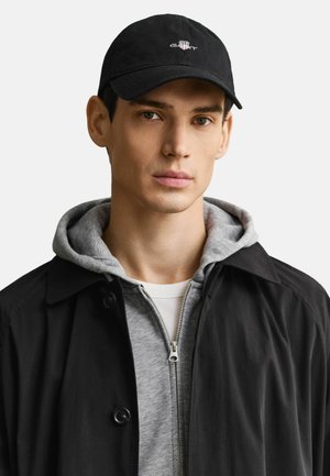 Joven con gorra de béisbol negra de GANT, sudadera gris con cremallera, camiseta blanca y abrigo negro, mirando hacia adelante.