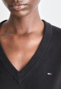 Pulover negru cu decolteu în V, cu guler ribat. Prezintă un mic logo cu accente roșu, alb și albastru pe piept. Textură moale, design ajustat.