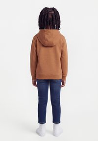 Παιδί με πλεξούδες που φοράει καφέ φούτερ με κουκούλα, μπλε τζιν, λευκές κάλτσες και λευκά παπούτσια, όρθιο με πλάτη προς το μέρος μας.