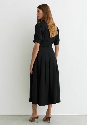 Femeie cu păr lung purtând o rochie midi neagră cu mâneci scurte și sandale maro cu toc, stând în picioare lângă un perete alb simplu.