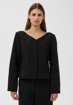 Femme aux longs cheveux bruns porte un blazer noir à col en V et un pantalon assorti, debout devant un fond clair et uni.