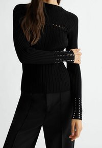 Pull en maille côtelée noire à manches longues, avec des découpes et des ornements le long des poignets. Coupe ajustée et cintrée à la taille.