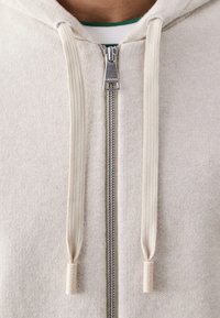 Šviesiai smėlio spalvos zip-up džemperis su sidabriniu užtrauktuku, grietinėlės spalvos raišteliais ir mažuoju Lacoste logotipu ant raištelių galų. Minkšta tekstūra ir kasdienis dizainas.
