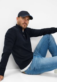 Maglione navy a zip collo alto, jeans blu e un berretto scuro. Il maglione ha polsini e colletto a costine; i jeans sono a gamba dritta con una leggera lavaggio.