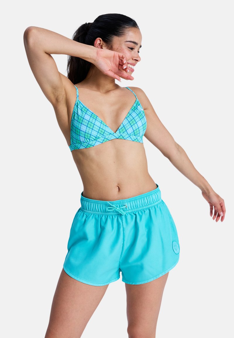 Młoda kobieta uśmiechająca się z uniesioną ręką, ubrana w niebieski kraciasty top bikini i turkusowe, jednolite szorty kąpielowe na białym tle.