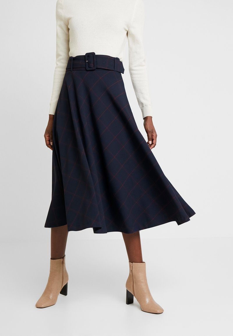 Jupe écossaise bleu marine avec une taille ceinturée, longueur au genou et tissu doux. Portée avec des bottines beiges à talon épais et fermeture éclair.