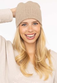 Beige knit beanie with ribbed texture, fitted shape, and seam detail. Model shows casual beige sweater with round neckline and frayed edges.