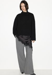 Schwarzer, übergroßer Pullover mit hohem Kragen; kombiniert mit weiten grauen Hosen mit schwarzem Satinbesatz und Spitzenbesatz.