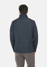 Giacca imbottita blu navy con colletto alto, toppe scure a contrasto sui gomiti e finitura liscia e texturizzata. Vista posteriore.