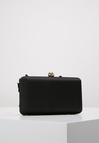 Clutch bag rettangolare nero con angoli arrotondati e una chiusura sferica dorata, posata su una superficie bianca su uno sfondo semplice.