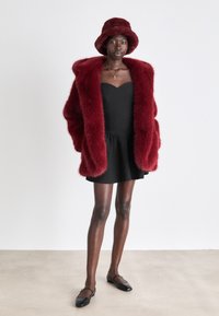 Burgunderfarget fuskepelsjakke i oversized stil, svart tettsittende kjole med volangkant, og en matchende burgunderfarget bøttehatt. Svart flate sko fullfører antrekket.
