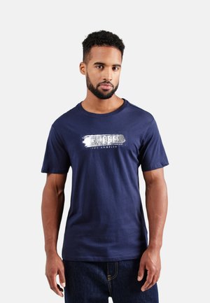 Camiseta de algodón azul marino con mangas cortas y cuello redondo. Presenta un logotipo gráfico plateado "GUESS LOS ÁNGELES" en la parte frontal.