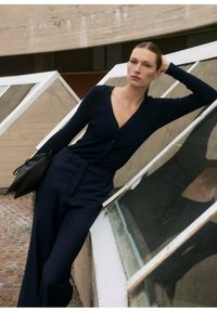 Cardigan navy a costine con scollo a V e chiusura a bottoni, abbinato a pantaloni navy a vita alta. Una borsa nera è posizionata sulla superficie.