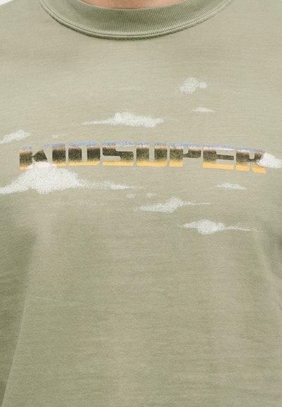Χακί βαμβακερό t-shirt με υφή, πολύχρωφο γραφικό που αναγράφει "KIDSUPER" με μοτίβα σύννεφων σε λευκό και ανοιχτό γκρι.