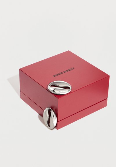 Κόκκινο κουτί κοσμημάτων Hugo Kreit με δύο λαμπερά ασημένια αφηρημένα οβάλ σκουλαρίκια τοποθετημένα πάνω και δίπλα του σε λευκό φόντο.