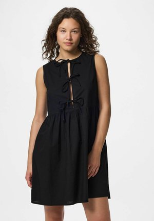 Jeune femme aux cheveux bouclés portant une robe noire sans manches avec des liens noués à l'avant et une taille froncée, se tenant devant un fond uni.