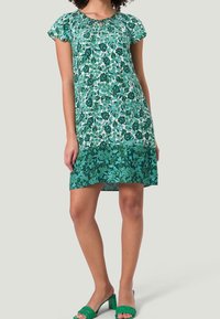 Robe à imprimé floral vert avec des manches courtes, un col froncé et un ourlet à volants. Portée avec des sandales tressées vertes.