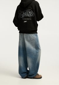Czarna bawełniana bluza z kapturem z nadrukiem na plecach, zestawiona z jasnoniebieskimi, luźnymi jeansami o wyblakłej fakturze oraz beżowymi butami trekkingowymi.