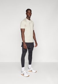 Beige idrettsgenser med logo, sammen med svarte shorts og tights. Hvite joggesko med svarte detaljer og teksturert såle.