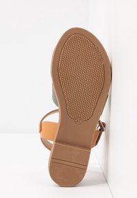Semelle de sandale marron avec une semelle texturée antidérapante, taille 37 indiquée, brides beiges et une boucle, placée debout contre un mur d'angle blanc.