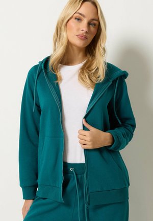 Vrouw draagt een tealgekleurde hoodie met rits over een wit shirt en bijpassende tealgekleurde joggingbroek tegen een effen achtergrond.