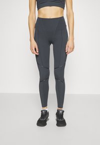 Spodnie legginsy w kolorze szarym z wysokim stanem, z kontrastowymi szwami, dopasowanym krojem i materiałem odprowadzającym wilgoć, w połączeniu z czarnymi butami sportowymi.