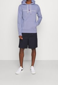 Felpa viola chiaro con logo bianco "Tommy Hilfiger", tasca a marsupio, cordini. Abbinata a pantaloni corti scuri e sneaker bianche.