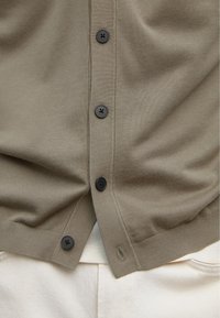 Gros plan d'un cardigan beige à boutons sur un pantalon blanc, montrant quatre boutons noirs et la texture en tissu tricoté.