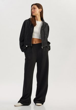 Ung kvinde med langt brunt hår iført sort flettet lynlås-hættetrøje, hvid cropped top, løse sorte bukser og hvide sneakers, stående med hænderne i lommerne.