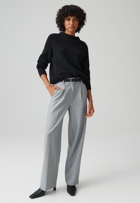 Maglione nero a coste abbinato a pantaloni a gamba larga grigio chiaro, con una cintura nera e stivali neri con tacco a punta, su uno sfondo neutro.