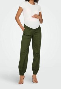 Zielone spodnie cargo z elastycznymi mankietami, z bocznymi kieszeniami i luźnym krojem. Modelka ma na sobie białą prążkowaną koszulkę i szpilki.