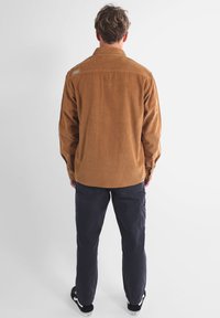 Chemise en velours côtelé marron à coupe droite, manches longues et col à boutons. Associée à un pantalon foncé et des baskets noires.