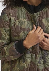 Ręce zapinają guziki na zielono-brązowej kurtce w kamuflaż, pikowanej, z czarnymi ściągaczami u mankietów i kołnierza.