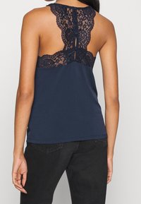 Tmavě modrý top na ramínka s krajkovým detailem na zádech, který zahrnuje výřez ve tvaru klíčové dírky a knoflíky. Měkká látka s přiléhavým střihem a zaobleným lemem.