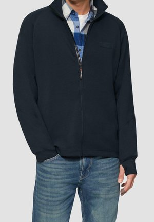 Mand iført blå lynlåstrøje over blå ternet skjorte og blå jeans, står med en hånd i lommen foran en ensfarvet baggrund.