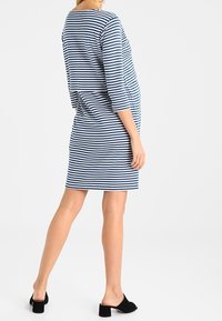 Kvinna iklädd en blå- och vitrandig knälång klänning med trekvartslånga ärmar och svarta blockklackade slip-on skor, stående på sidan.