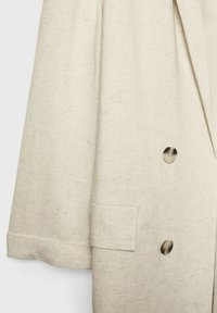 Gros plan d'une veste en lin beige montrant deux boutons, une poche avec rabat et une partie de manche avec un poignet.