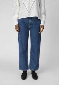 Kvinna i vit blus med långa ärmar och broderade detaljer, blå raka jeans, svarta strumpbyxor och svarta Mary Jane-skor, ståendes upprätt.