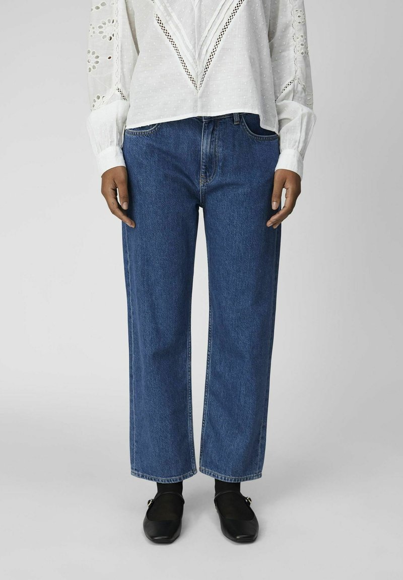 Kvinna i vit blus med långa ärmar och broderade detaljer, blå raka jeans, svarta strumpbyxor och svarta Mary Jane-skor, ståendes upprätt.