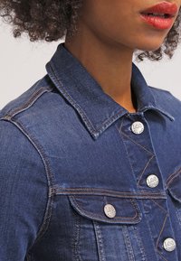 Mörkblå denimjacka med krage, silverknappar och bröstfickor. Har kontrasterande sömmar och en figurnära design.