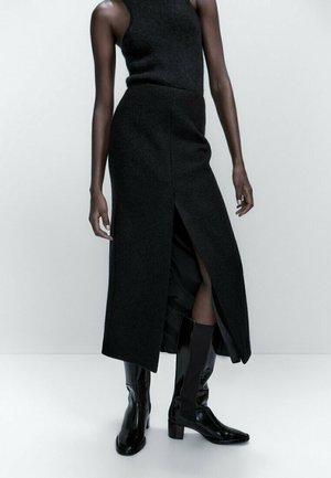Sleeveless black midi dress with front slit, worn with black glossy mid-calf heeled boots on a standing model against plain background.